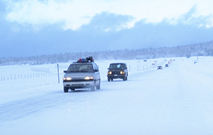 Photo.  A line of cars is shown traveling on a snowy road. 