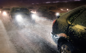 Photo.  Cars with lights on on a very snowy road with the snow coming down.  The ice and snow is visible on the road.