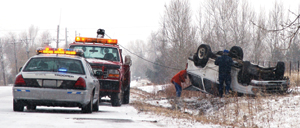 Photo.  A police car and a two truck are shown alongside an SUV that has left the roadway and overturned in an area next to the road.  Two individuals are shown working on the crashed car.