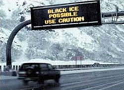 This photo shows a car on a snowy, ice road with a Digital Message Sign above saying Black Ice Possible, Use Caution. The road is an area with mountains that area shown to the left of the roadway.