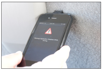 Photo of a smart phone showing a red triangle with an exclamation point in the middle of it and the words 'Warning! Application Disabled While Driving.'