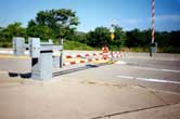 Photograph - Cantaliever type autmated gates.  One gate postioned in the foreground is in the "closed" position.  The other gate is in the "up" position and directly accross the roadway from the closed gate.