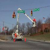 Photograph:    Two workers are perfoming maintenace on a traffic signal.  One worker is standing near the rear end of a bucket truck, while the other worker is standing in the bucket, which has been lifted just below the signal head.  