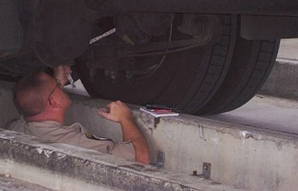 Figure 6. Photograph. Inspector standing in inspection bay beneath a semi tractor-trailer, using a ruler to measure the brake stroke of a truck in the inspection shed during a routine inspection at Laurel County, Kentucky.